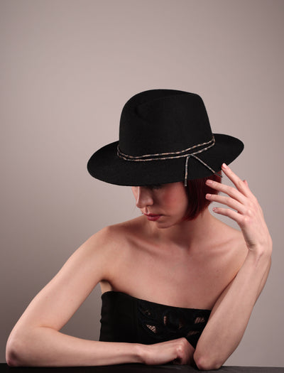 The model showcases the left high angle of the Stella fedora crafted in deep black felt, the silhouette is elevated by a subtle metallic crystal band featuring strands of crystal embellished thread that catch the light with every tilt of the head. The tied ends fall just off-centre on the left side. Designed by Merve Bayindir.