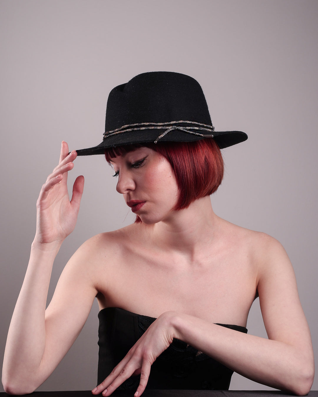 The model showcases the left side of the Stella fedora crafted in deep black felt, the silhouette is elevated by a subtle metallic crystal band featuring strands of crystal embellished thread that catch the light with every tilt of the head. The tied ends fall just off-centre on the left side. Designed by Merve Bayindir.
