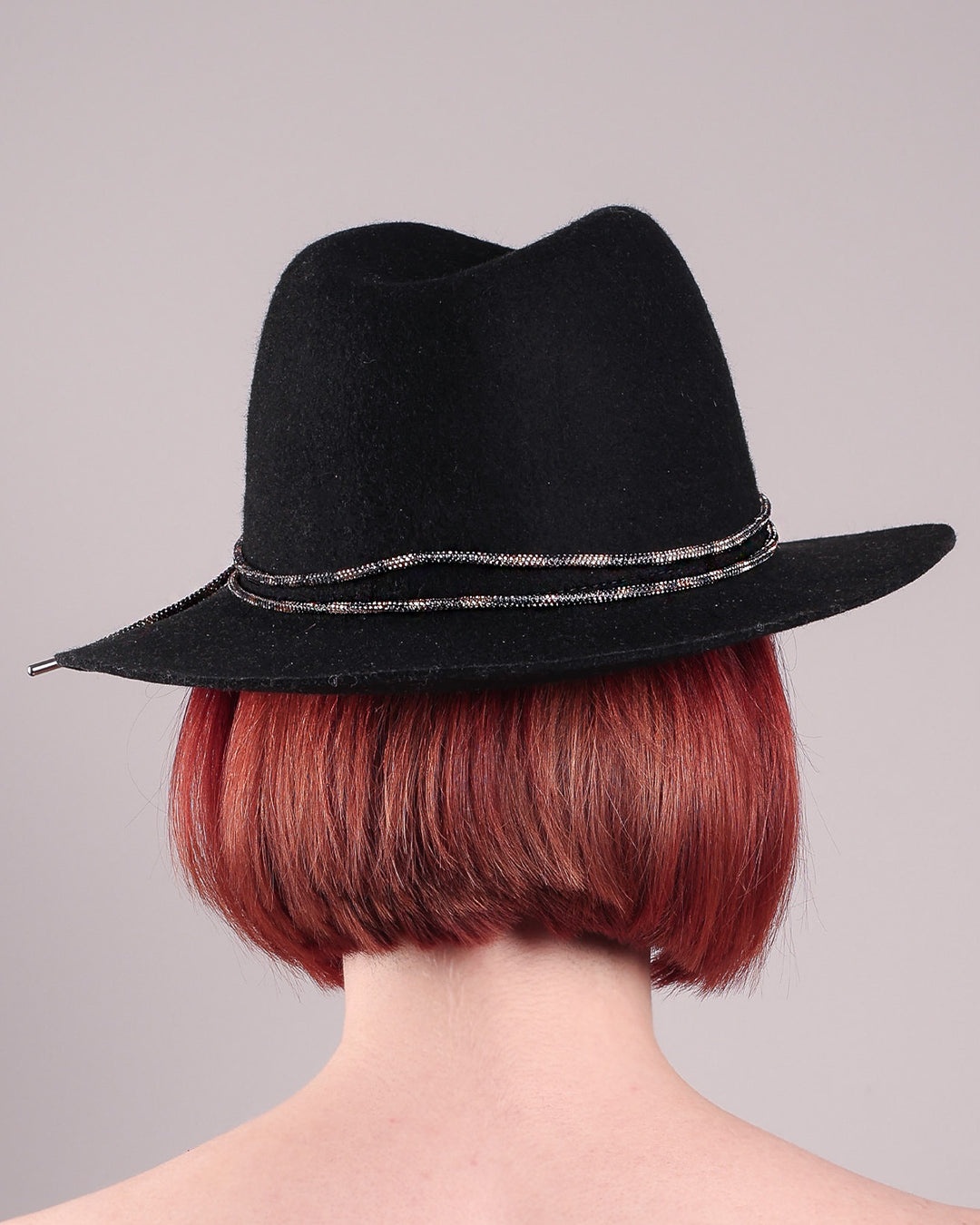 The model showcases the back of the Stella fedora crafted in deep black felt, the silhouette is elevated by a subtle metallic crystal band featuring strands of crystal embellished thread that catch the light with every tilt of the head. The tied ends fall just off-centre on the left side. Designed by Merve Bayindir.