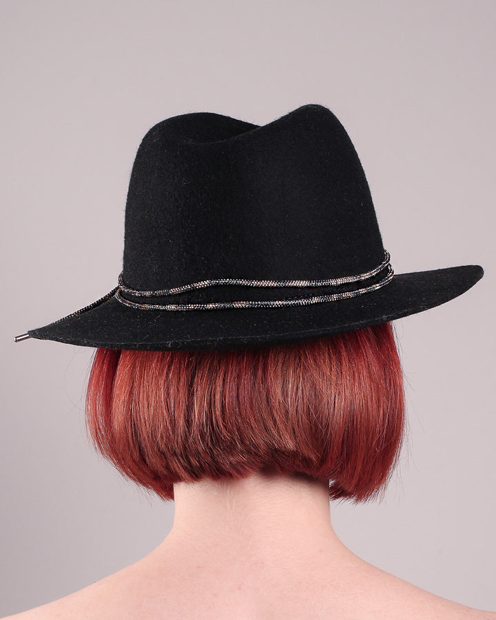 The model showcases the back of the Stella fedora crafted in deep black felt, the silhouette is elevated by a subtle metallic crystal band featuring strands of crystal embellished thread that catch the light with every tilt of the head. The tied ends fall just off-centre on the left side. Designed by Merve Bayindir.