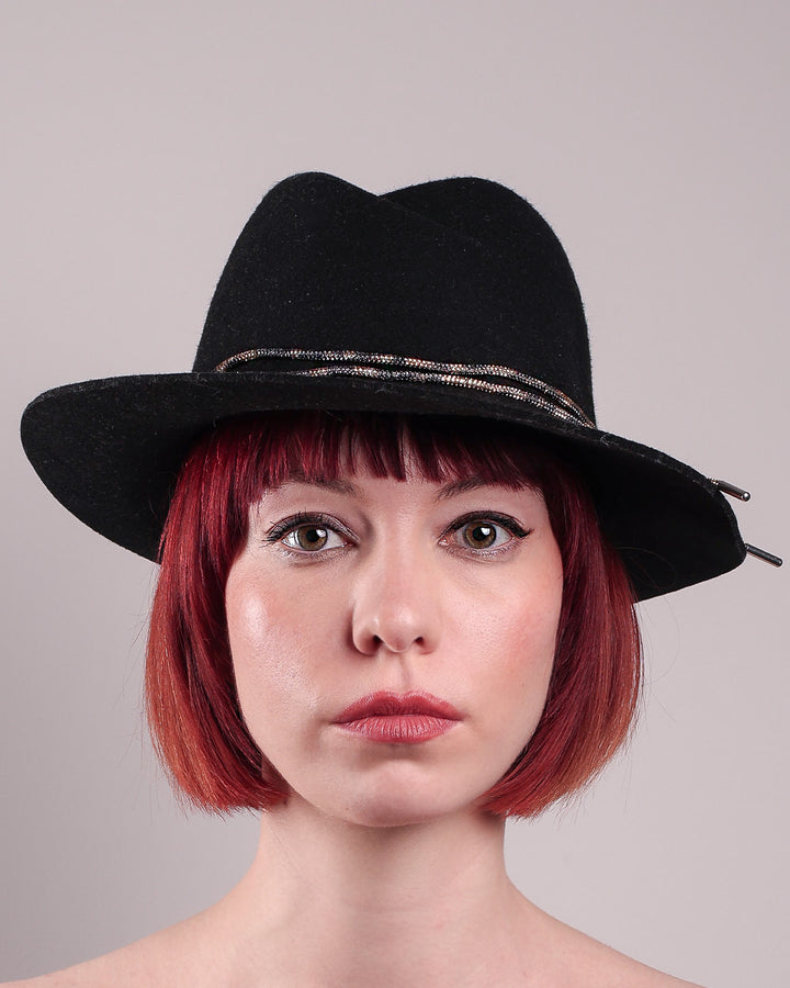 The model showcases the front close up of the Stella fedora crafted in deep black felt, the silhouette is elevated by a subtle metallic crystal band featuring strands of crystal embellished thread that catch the light with every tilt of the head. The tied ends fall just off-centre on the left side. Designed by Merve Bayindir.