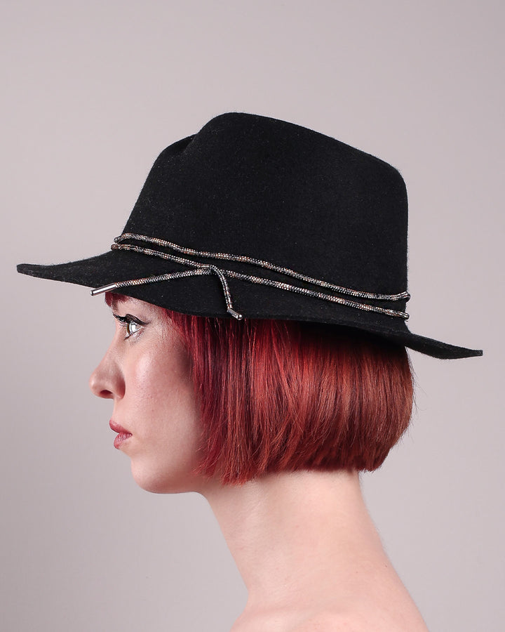 The model showcases the left side close up of the Stella fedora crafted in deep black felt, the silhouette is elevated by a subtle metallic crystal band featuring strands of crystal embellished thread that catch the light with every tilt of the head. The tied ends fall just off-centre on the left side. Designed by Merve Bayindir.