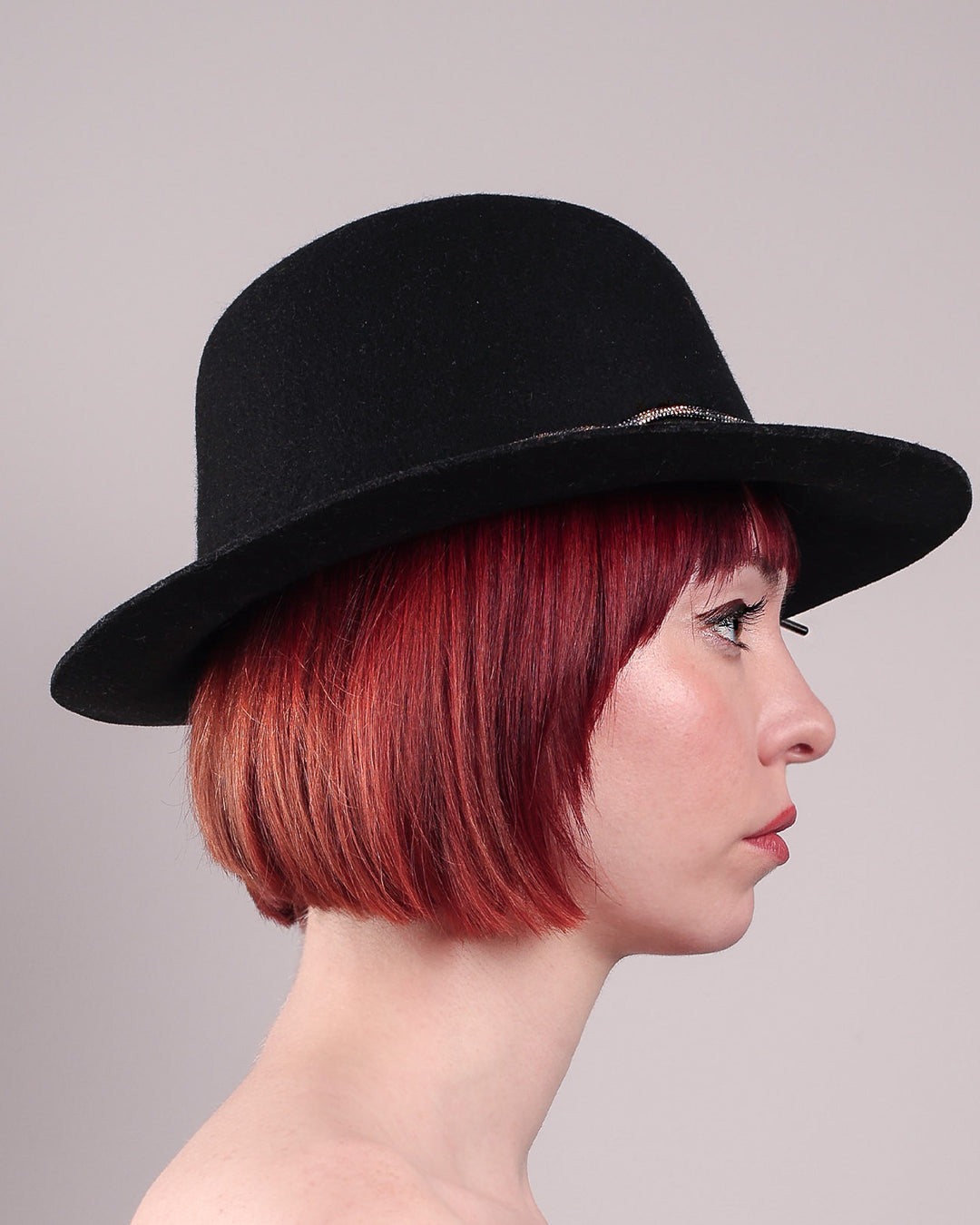 The model showcases the right side close up of the Stella fedora crafted in deep black felt, the silhouette is elevated by a subtle metallic crystal band featuring strands of crystal embellished thread that catch the light with every tilt of the head. The tied ends fall just off-centre on the left side. Designed by Merve Bayindir.