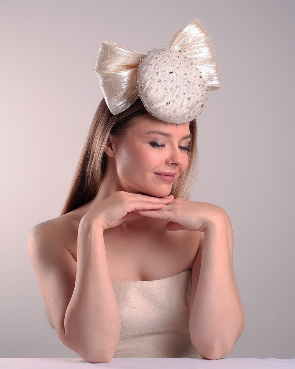 Model is showcasing the right side front view of the grey double button fascinator is worn to the right front of the head. The high back is decorated with silver shimmering large bow while the front is detailed with different sized crystals. Fitted with a wire headband and elastic