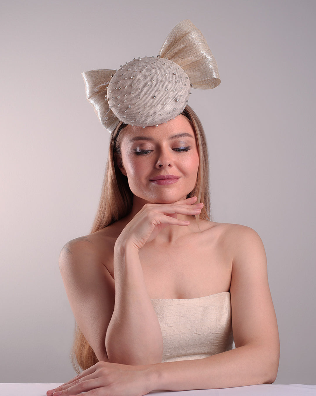 Model is showcasing the front view of the grey double button fascinator is worn to the right front of the head. The high back is decorated with silver shimmering large bow while the front is detailed with different sized crystals. Fitted with a wire headband and elastic