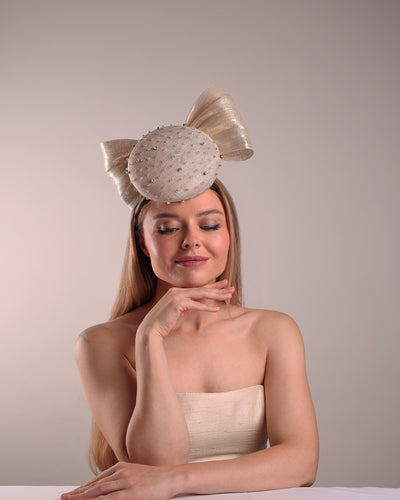 Model is showcasing the front view of the grey double button fascinator is worn to the right front of the head. The high back is decorated with silver shimmering large bow while the front is detailed with different sized crystals. Fitted with a wire headband and elastic