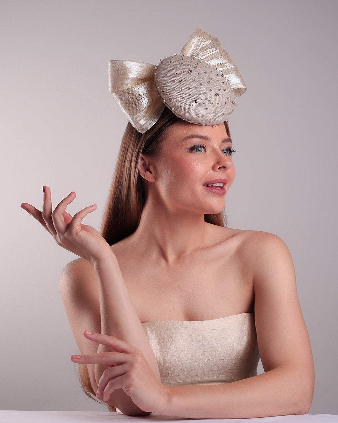 Model is showcasing the right front view from the below of the grey double button fascinator is worn to the right front of the head. The high back is decorated with silver shimmering large bow while the front is detailed with different sized crystals. Fitted with a wire headband and elastic