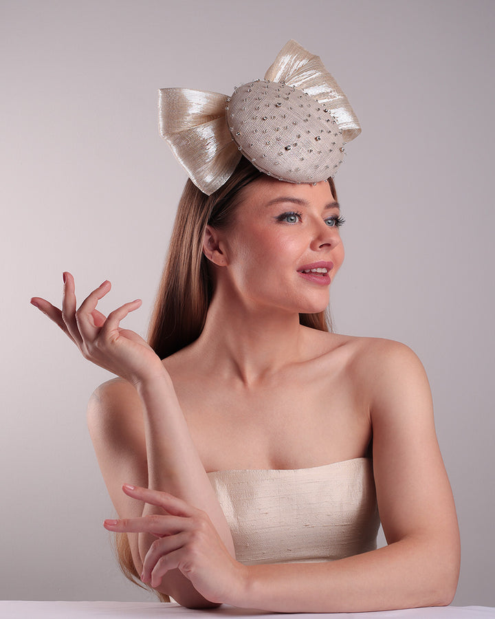 Model is showcasing the right front view from the below of the grey double button fascinator is worn to the right front of the head. The high back is decorated with silver shimmering large bow while the front is detailed with different sized crystals. Fitted with a wire headband and elastic