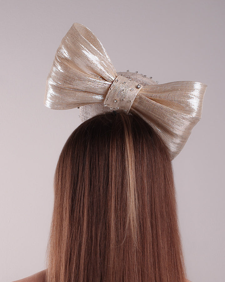 Model is showcasing the back view of the grey double button fascinator is worn to the right front of the head. The high back is decorated with silver shimmering large bow while the front is detailed with different sized crystals. Fitted with a wire headband and elastic
