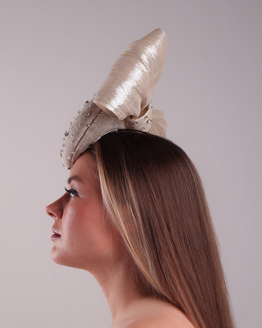 Model is showcasing the left side view of the grey double button fascinator is worn to the right front of the head. The high back is decorated with silver shimmering large bow while the front is detailed with different sized crystals. Fitted with a wire headband and elastic