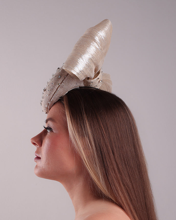 Model is showcasing the left side view of the grey double button fascinator is worn to the right front of the head. The high back is decorated with silver shimmering large bow while the front is detailed with different sized crystals. Fitted with a wire headband and elastic