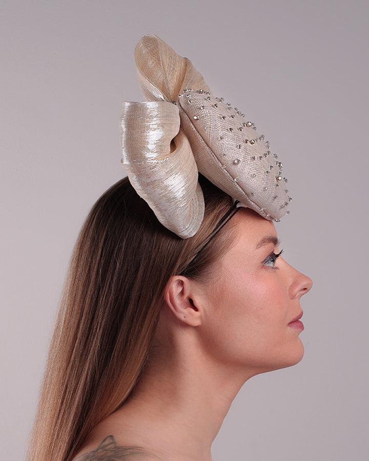 Model is showcasing the right side view of the grey double button fascinator is worn to the right front of the head. The high back is decorated with silver shimmering large bow while the front is detailed with different sized crystals. Fitted with a wire headband and elastic