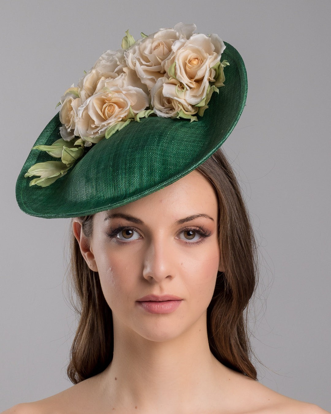 Model is showcasing the front side close up of medium sized emerald green saucer hat decorated with a bouquet on top back side. The bouquet features six flowers in different sizes and shades ranging from light peach to yellow. Designed and made by Merve Bayindir Millinery.