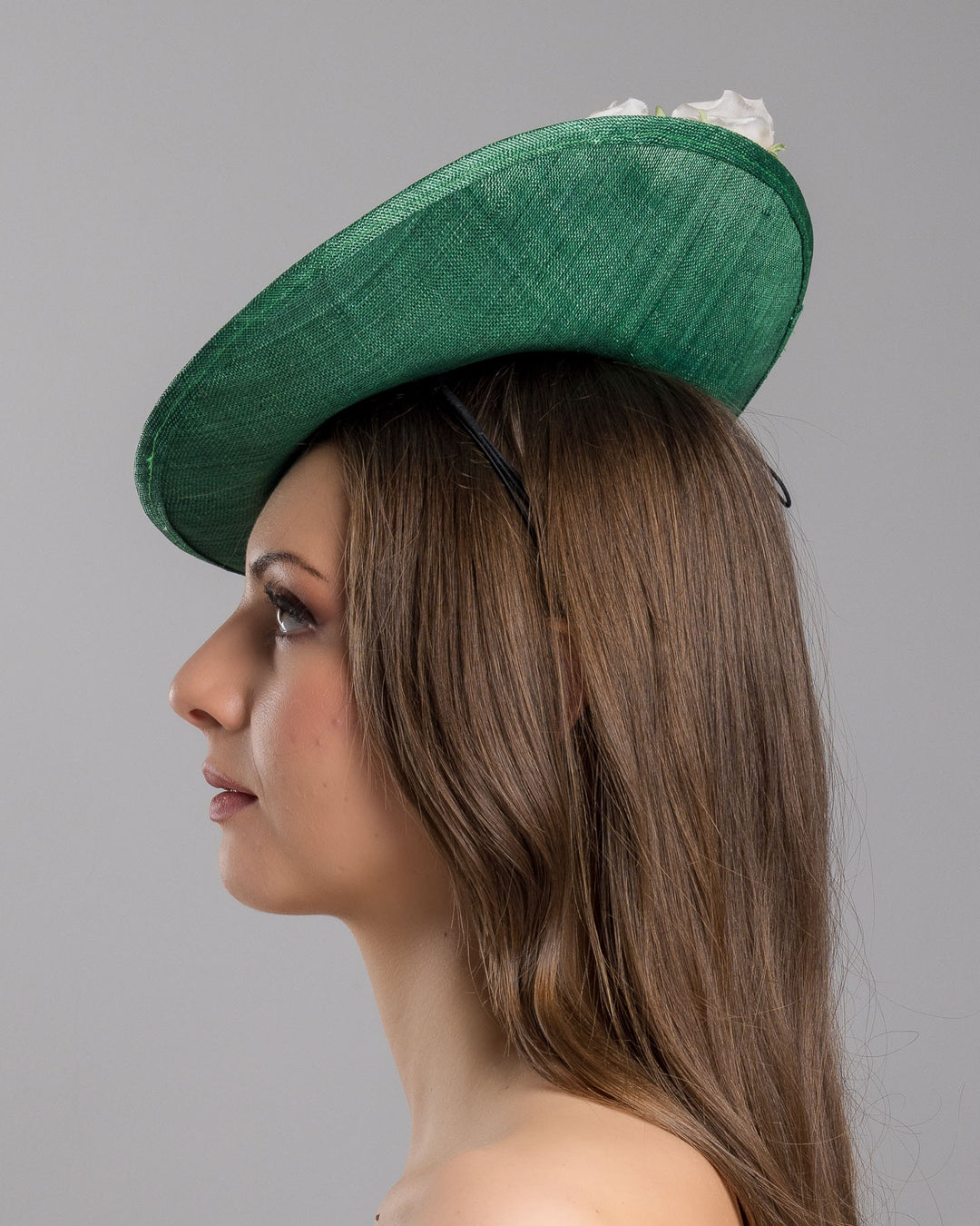 Model is showcasing the left side close up of medium sized emerald green saucer hat decorated with a bouquet on top back side. The bouquet features six flowers in different sizes and shades ranging from light peach to yellow. Designed and made by Merve Bayindir Millinery.