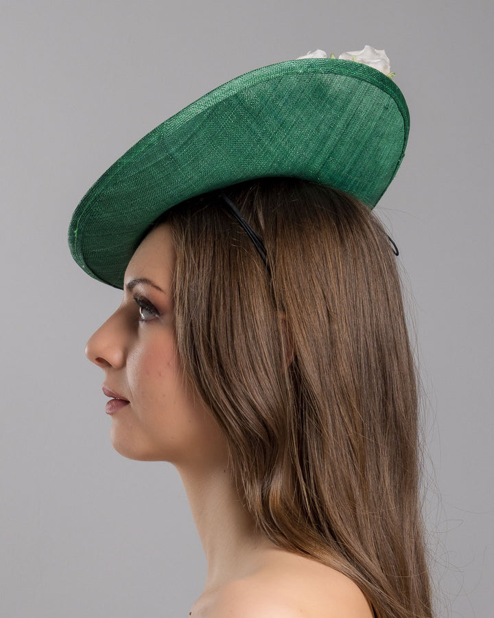 Model is showcasing the left side close up of medium sized emerald green saucer hat decorated with a bouquet on top back side. The bouquet features six flowers in different sizes and shades ranging from light peach to yellow. Designed and made by Merve Bayindir Millinery.