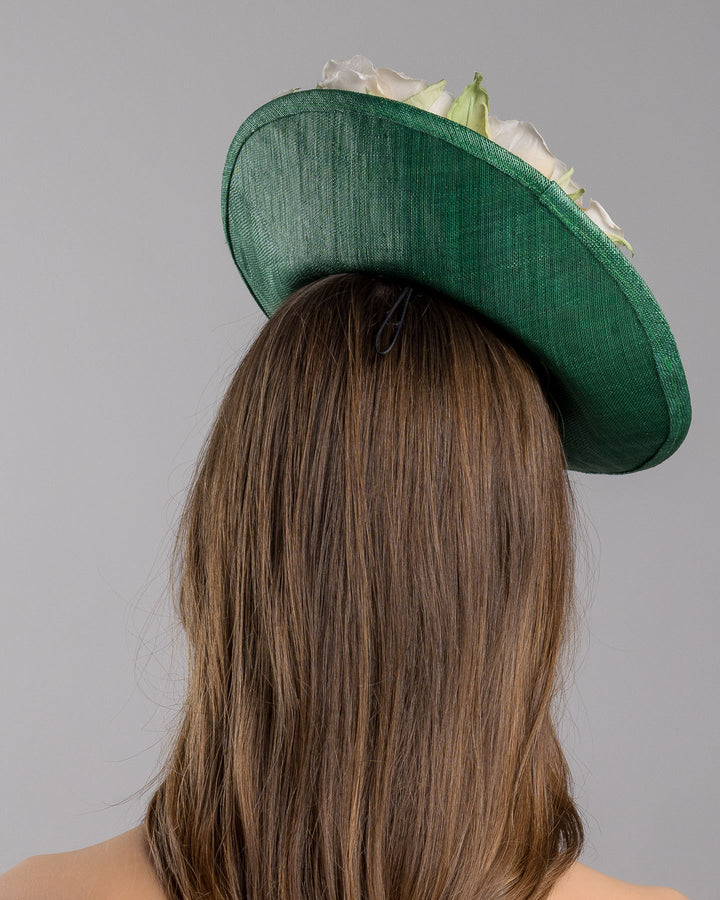 Model is showcasing the back side close up of medium sized emerald green saucer hat decorated with a bouquet on top back side. The bouquet features six flowers in different sizes and shades ranging from light peach to yellow. Designed and made by Merve Bayindir Millinery.