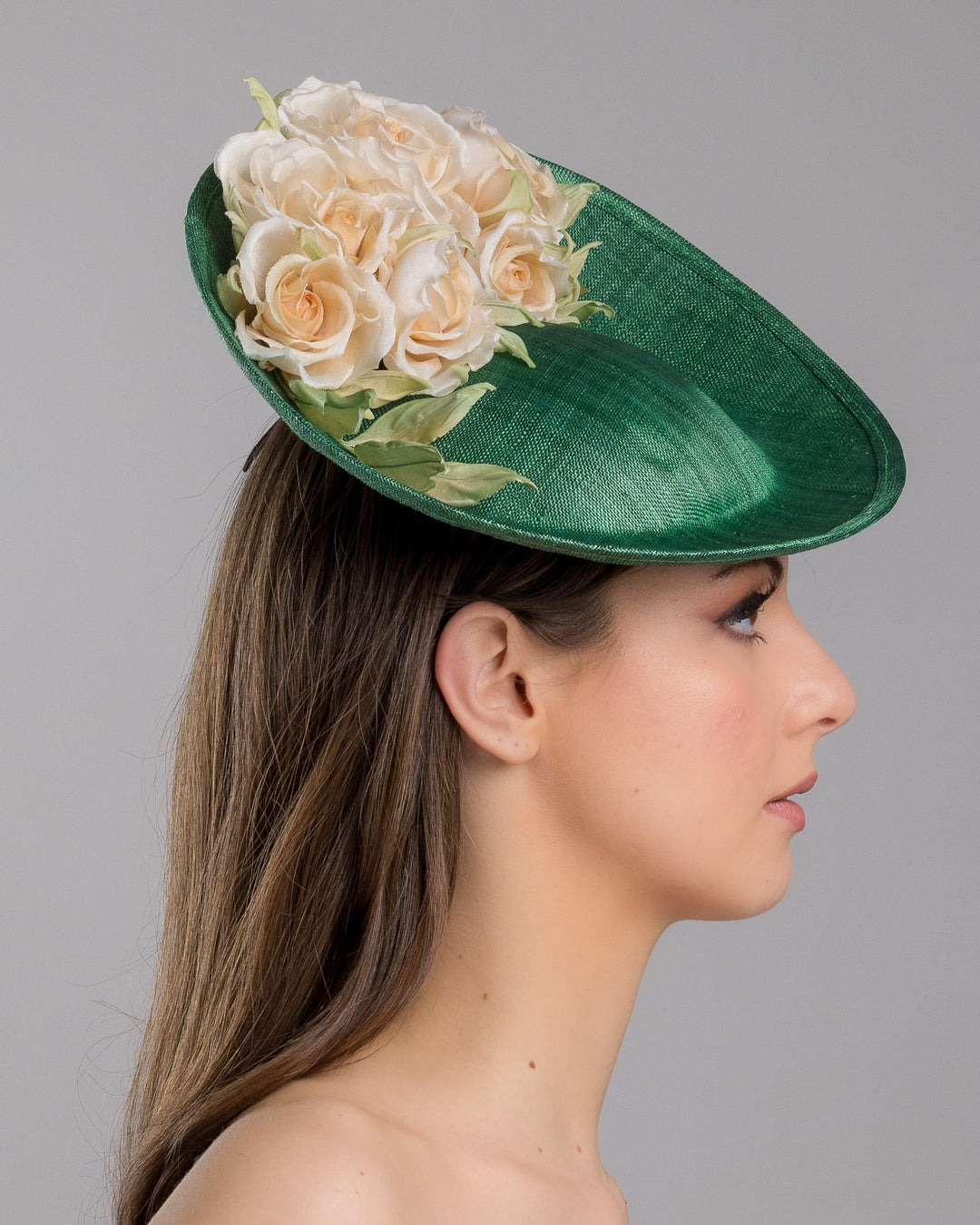Model is showcasing the right side close up of medium sized emerald green saucer hat decorated with a bouquet on top back side. The bouquet features six flowers in different sizes and shades ranging from light peach to yellow. Designed and made by Merve Bayindir Millinery.