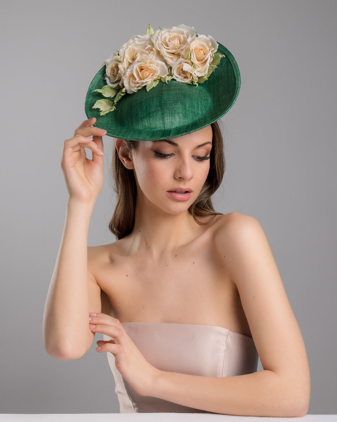 Female model is posing wearing Stevie medium sized emerald green saucer hat decorated with a bouquet on top back side. The bouquet features six flowers in different sizes and shades ranging from light peach to yellow. Designed and made by Merve Bayindir Millinery.
