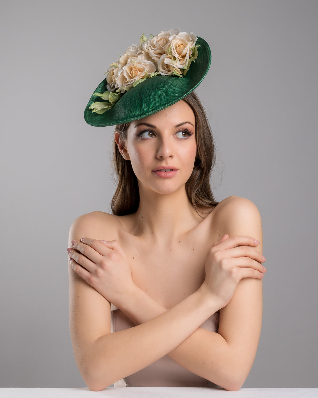 Female model is posing wearing Stevie medium sized emerald green saucer hat decorated with a bouquet on top back side. The bouquet features six flowers in different sizes and shades ranging from light peach to yellow. Designed and made by Merve Bayindir Millinery.