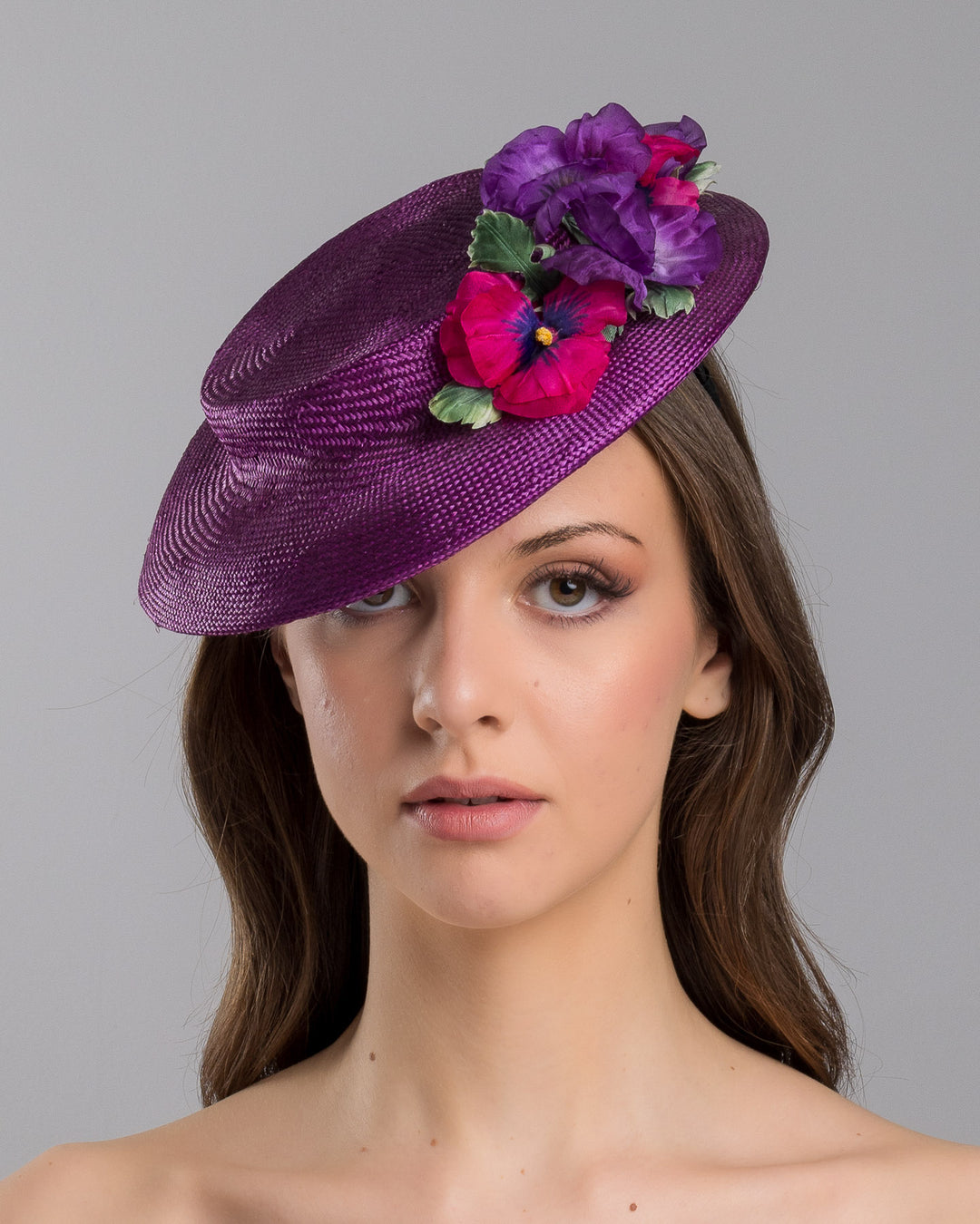The model is showcasing the front side close up of the purple parasisal mini boater hat decorated with purple and hot pink violets on the left side. Worn to the right side of the head. Designed and made by Merve Bayindir Millinery.