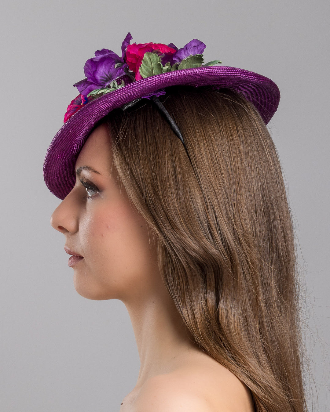 The model is showcasing the left side close up of the purple parasisal mini boater hat decorated with purple and hot pink violets on the left side. Worn to the right side of the head. Designed and made by Merve Bayindir Millinery.