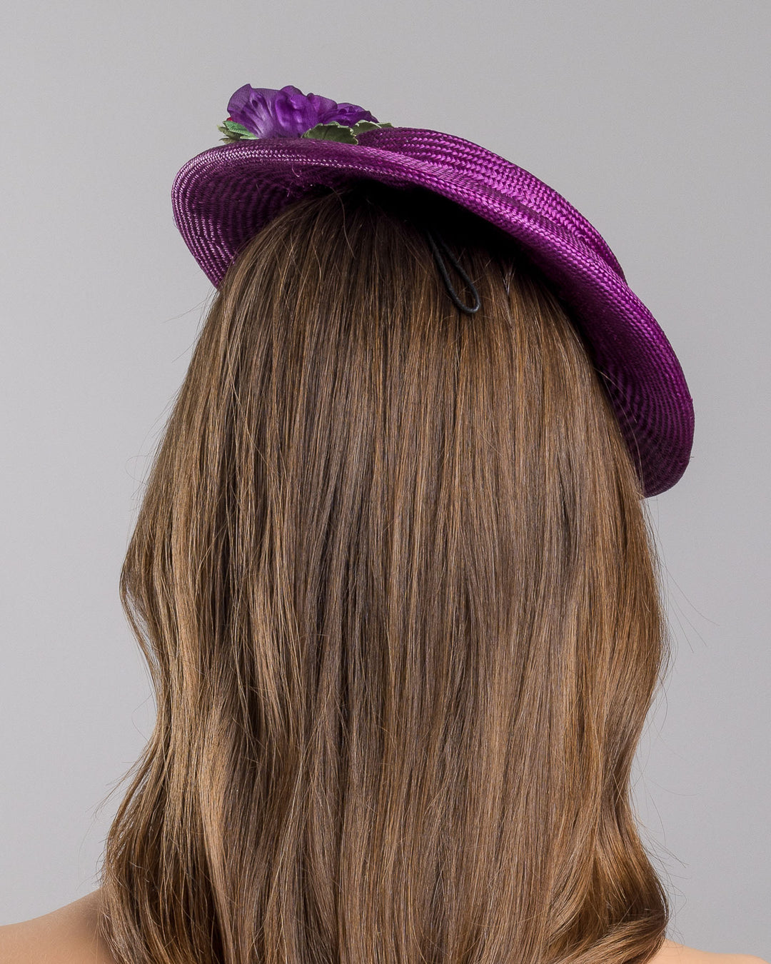 The model is showcasing the back side close up of the purple parasisal mini boater hat decorated with purple and hot pink violets on the left side. Worn to the right side of the head. Designed and made by Merve Bayindir Millinery.