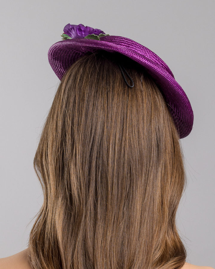 The model is showcasing the back side close up of the purple parasisal mini boater hat decorated with purple and hot pink violets on the left side. Worn to the right side of the head. Designed and made by Merve Bayindir Millinery.