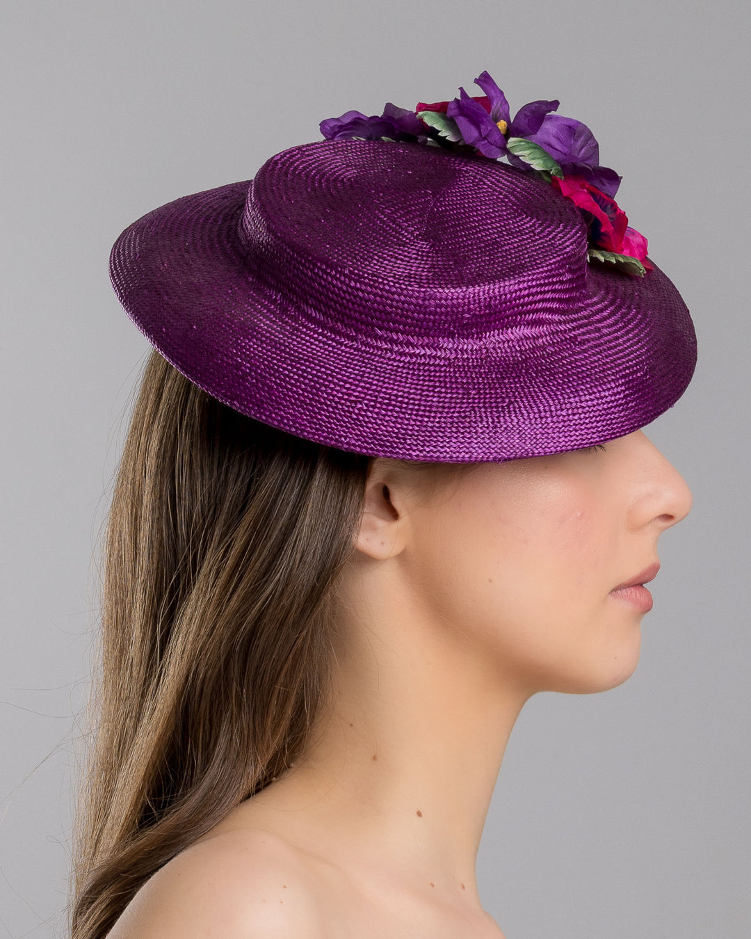 The model is showcasing the right side close up of the purple parasisal mini boater hat decorated with purple and hot pink violets on the left side. Worn to the right side of the head. Designed and made by Merve Bayindir Millinery.
