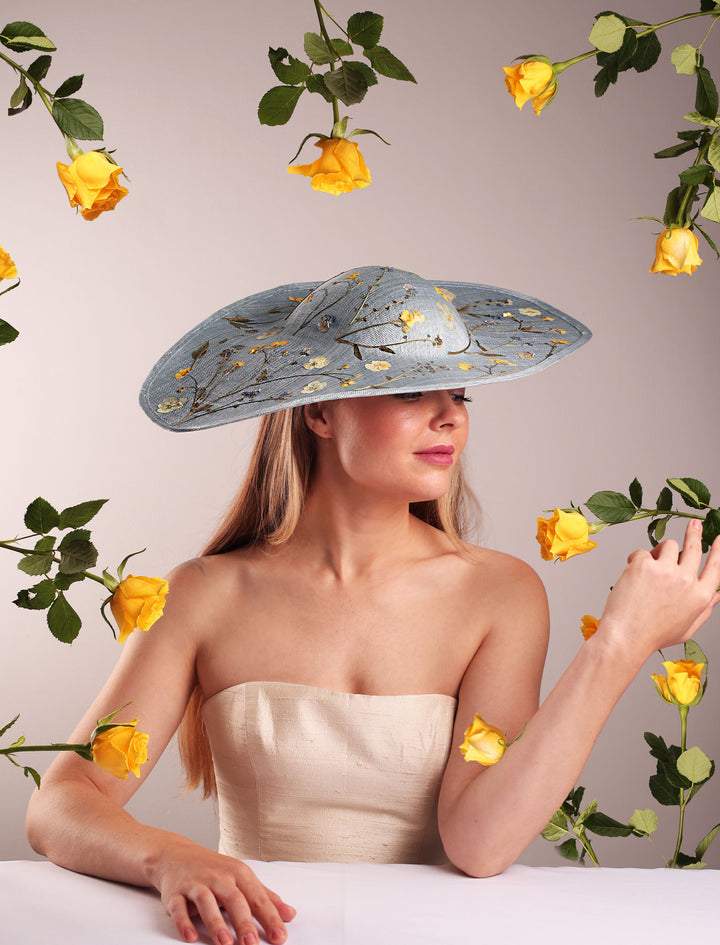 Woman wearing a decorative hat with flowers, surrounded by yellow roses on a light background