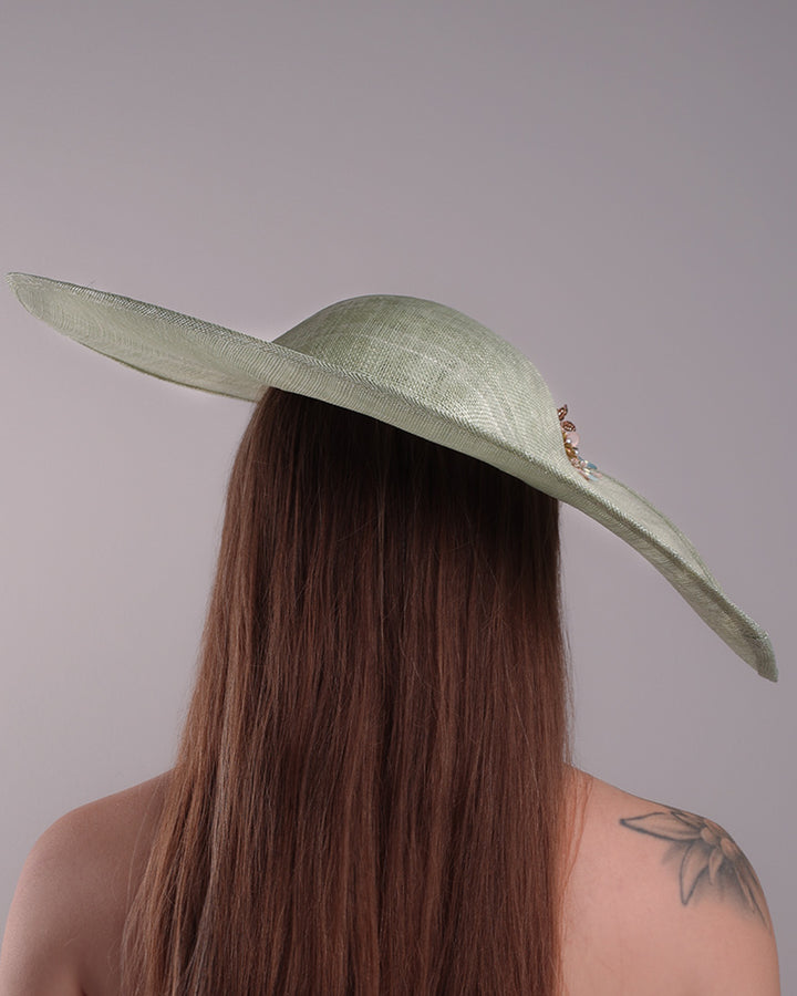 Model is showcasing the back side of green wide brim long hat, tilted to the right side swings upwards to the lefts side with a curve. Front top right side of the hat is decorated with multi colour resin flowers. Fitted to the head with wire headband and elastic.