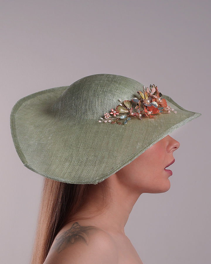 Model is showcasing the right side of green wide brim long hat, tilted to the right side swings upwards to the lefts side with a curve. Front top right side of the hat is decorated with multi colour resin flowers. Fitted to the head with wire headband and elastic.