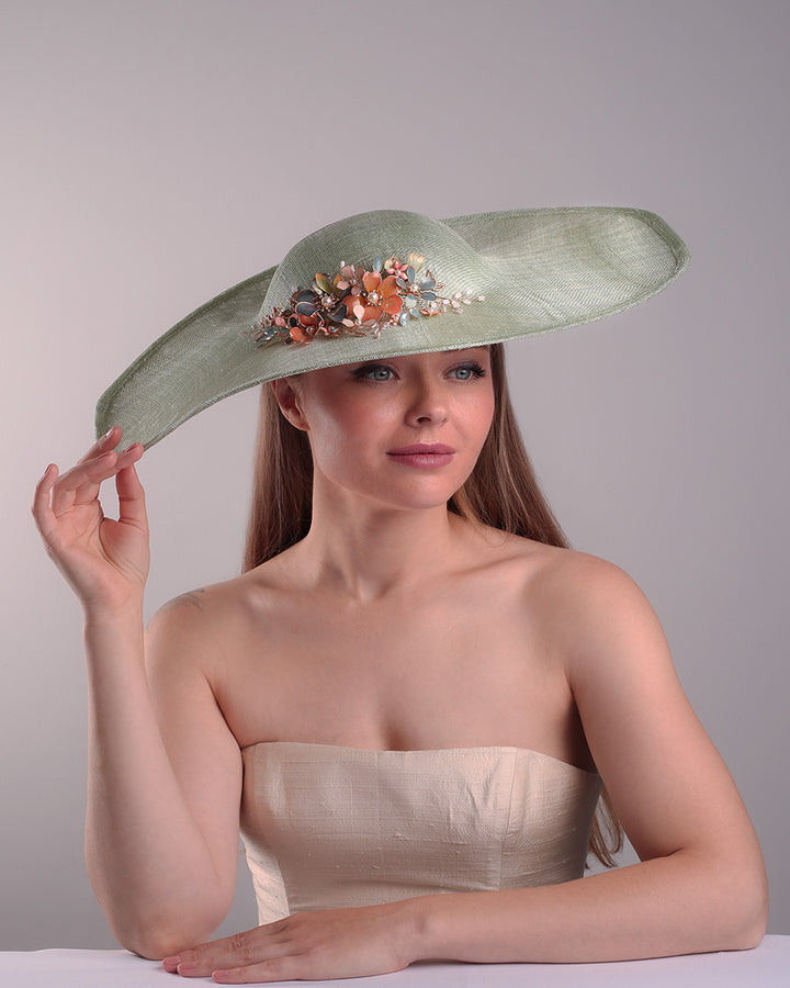 Model is showcasing the front side of green wide brim long hat, tilted to the right side swings upwards to the lefts side with a curve. Front top right side of the hat is decorated with multi colour resin flowers. Fitted to the head with wire headband and elastic.