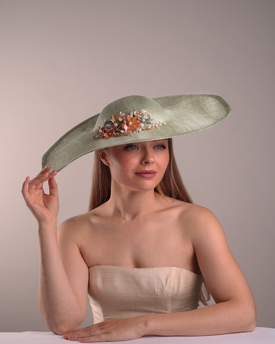 Model is showcasing the front side of green wide brim long hat, tilted to the right side swings upwards to the lefts side with a curve. Front top right side of the hat is decorated with multi colour resin flowers. Fitted to the head with wire headband and elastic.