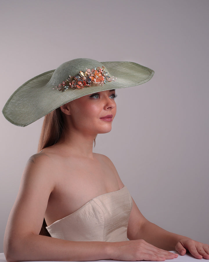 Model is showcasing the right front side of green wide brim long hat, tilted to the right side swings upwards to the lefts side with a curve. Front top right side of the hat is decorated with multi colour resin flowers. Fitted to the head with wire headband and elastic.