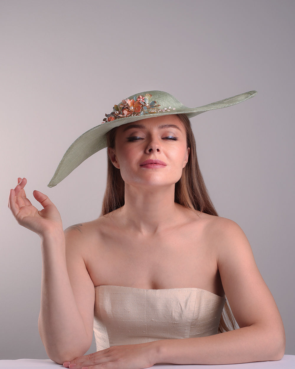 Model is showcasing the front side from the lower angle of green wide brim long hat, tilted to the right side swings upwards to the lefts side with a curve. Front top right side of the hat is decorated with multi colour resin flowers. Fitted to the head with wire headband and elastic.