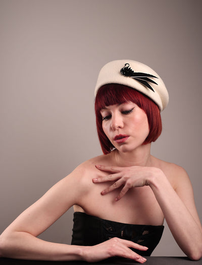 The model showcases the right front of the Swan Felt Beret Hat, crafted from soft white felt and structured to sit perched to the side. A sculpted black swan with rhinestones glides outward in layers, featuring fan-like leather feathers at the end, which elegantly contrast with the creamy white base. Designed by Merve Bayindir.