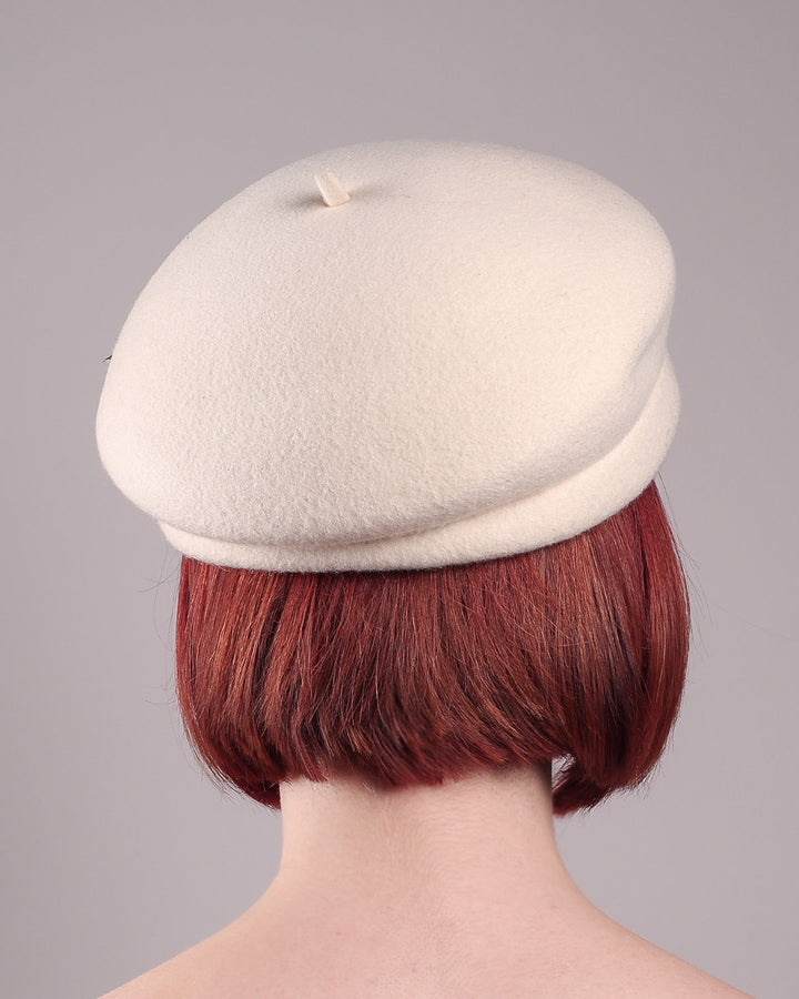 The model showcases the back of the Swan Felt Beret Hat, crafted from soft white felt and structured to sit perched to the side. A sculpted black swan with rhinestones glides outward in layers, featuring fan-like leather feathers at the end, which elegantly contrast with the creamy white base. Designed by Merve Bayindir.