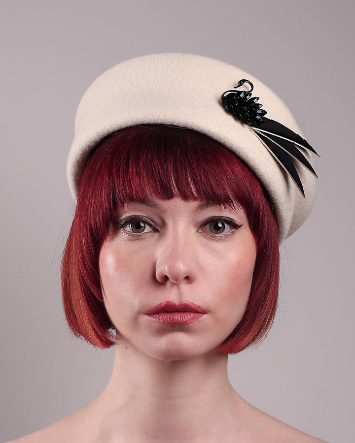 The model showcases the front close up of the Swan Felt Beret Hat, crafted from soft white felt and structured to sit perched to the side. A sculpted black swan with rhinestones glides outward in layers, featuring fan-like leather feathers at the end, which elegantly contrast with the creamy white base. Designed by Merve Bayindir.