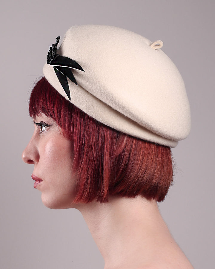 The model showcases the left side close up of the Swan Felt Beret Hat, crafted from soft white felt and structured to sit perched to the side. A sculpted black swan with rhinestones glides outward in layers, featuring fan-like leather feathers at the end, which elegantly contrast with the creamy white base. Designed by Merve Bayindir.