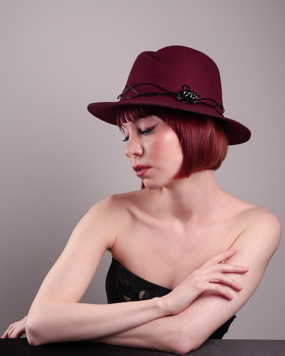 The model showcases the left side of the Sylph fedora hat, a refined statement in deep burgundy felt, structured with clean, classic lines and softened by an ethereal twist: a fluid black wire detail weaves around the crown like delicate ink on parchment, with a crystal-embellished black metal swan on the left side. The embellishment feels both organic and intentional—a subtle nod to surrealism, grounded in wearable form.  Designed by Merve Bayindir.