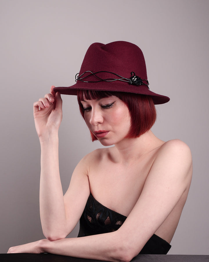 The model showcases the front left side of the Sylph fedora hat, a refined statement in deep burgundy felt, structured with clean, classic lines and softened by an ethereal twist: a fluid black wire detail weaves around the crown like delicate ink on parchment, with a crystal-embellished black metal swan on the left side. The embellishment feels both organic and intentional—a subtle nod to surrealism, grounded in wearable form.  Designed by Merve Bayindir.