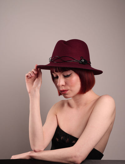 The model showcases the front left side of the Sylph fedora hat, a refined statement in deep burgundy felt, structured with clean, classic lines and softened by an ethereal twist: a fluid black wire detail weaves around the crown like delicate ink on parchment, with a crystal-embellished black metal swan on the left side. The embellishment feels both organic and intentional—a subtle nod to surrealism, grounded in wearable form.  Designed by Merve Bayindir.