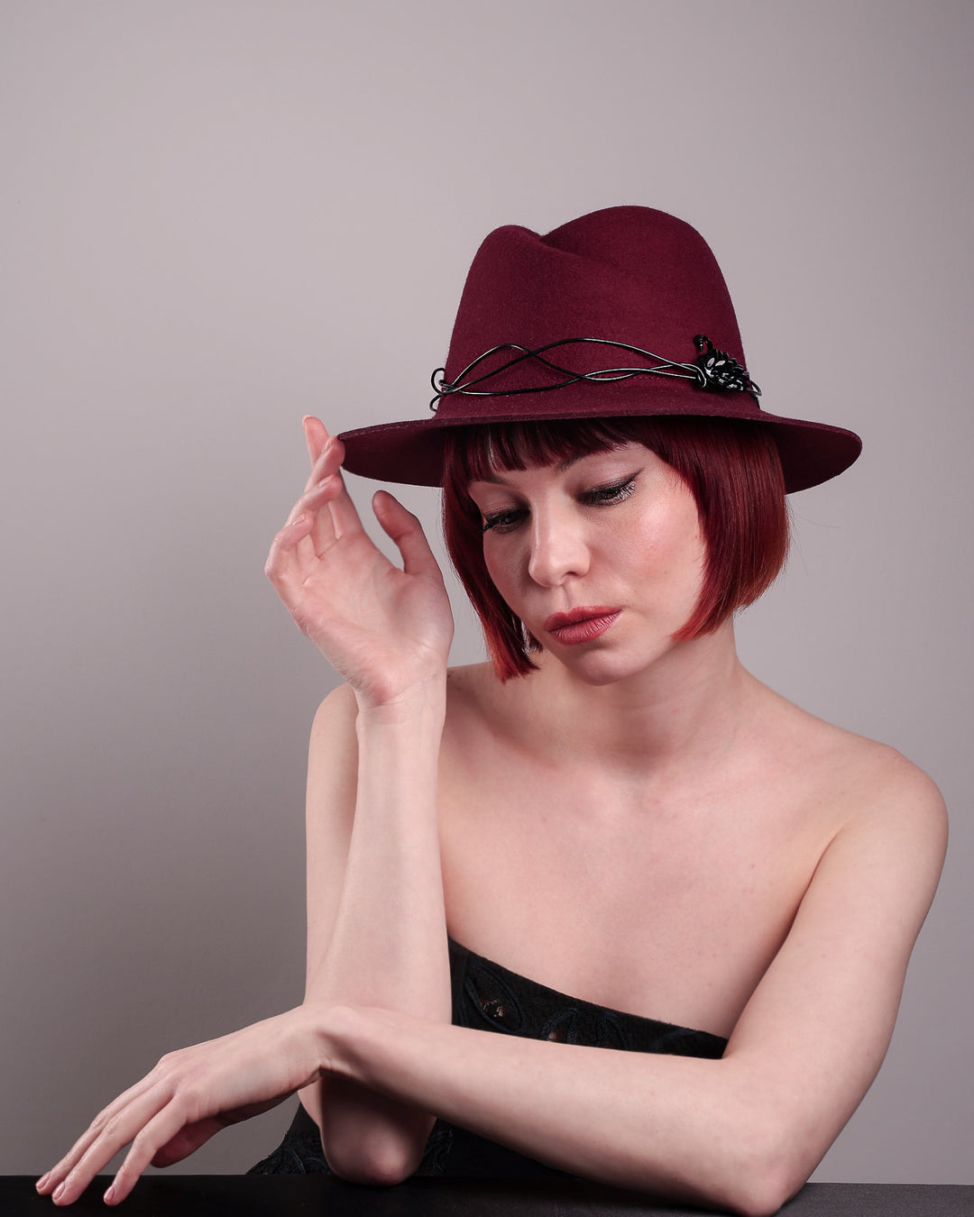 The model showcases the front left of the Sylph fedora hat, a refined statement in deep burgundy felt, structured with clean, classic lines and softened by an ethereal twist: a fluid black wire detail weaves around the crown like delicate ink on parchment, with a crystal-embellished black metal swan on the left side. The embellishment feels both organic and intentional—a subtle nod to surrealism, grounded in wearable form.  Designed by Merve Bayindir.