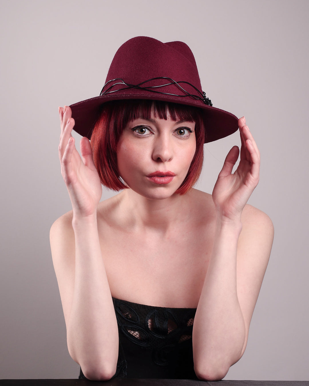 The model showcases the front of the Sylph fedora hat, a refined statement in deep burgundy felt, structured with clean, classic lines and softened by an ethereal twist: a fluid black wire detail weaves around the crown like delicate ink on parchment, with a crystal-embellished black metal swan on the left side. The embellishment feels both organic and intentional—a subtle nod to surrealism, grounded in wearable form.  Designed by Merve Bayindir.