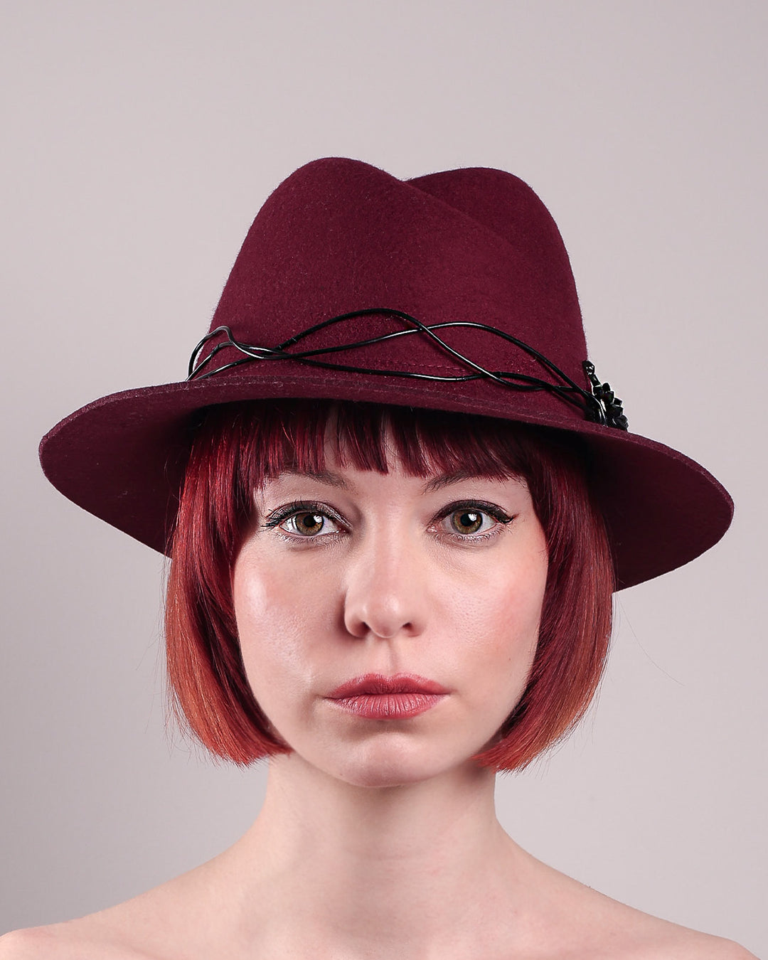 The model showcases the front close up of the Sylph fedora hat, a refined statement in deep burgundy felt, structured with clean, classic lines and softened by an ethereal twist: a fluid black wire detail weaves around the crown like delicate ink on parchment, with a crystal-embellished black metal swan on the left side. The embellishment feels both organic and intentional—a subtle nod to surrealism, grounded in wearable form.  Designed by Merve Bayindir.