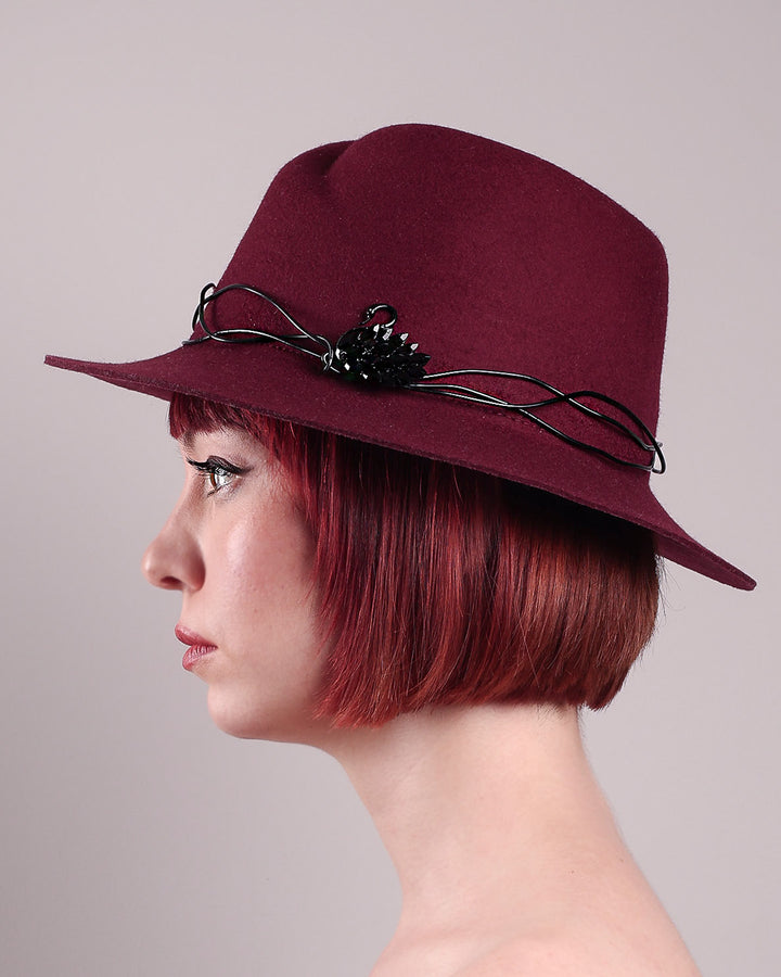 The model showcases the left side close up of the Sylph fedora hat, a refined statement in deep burgundy felt, structured with clean, classic lines and softened by an ethereal twist: a fluid black wire detail weaves around the crown like delicate ink on parchment, with a crystal-embellished black metal swan on the left side. The embellishment feels both organic and intentional—a subtle nod to surrealism, grounded in wearable form.  Designed by Merve Bayindir.