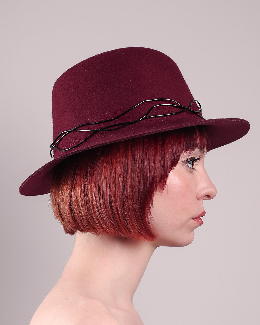 The model showcases the right side close up of the Sylph fedora hat, a refined statement in deep burgundy felt, structured with clean, classic lines and softened by an ethereal twist: a fluid black wire detail weaves around the crown like delicate ink on parchment, with a crystal-embellished black metal swan on the left side. The embellishment feels both organic and intentional—a subtle nod to surrealism, grounded in wearable form.  Designed by Merve Bayindir.