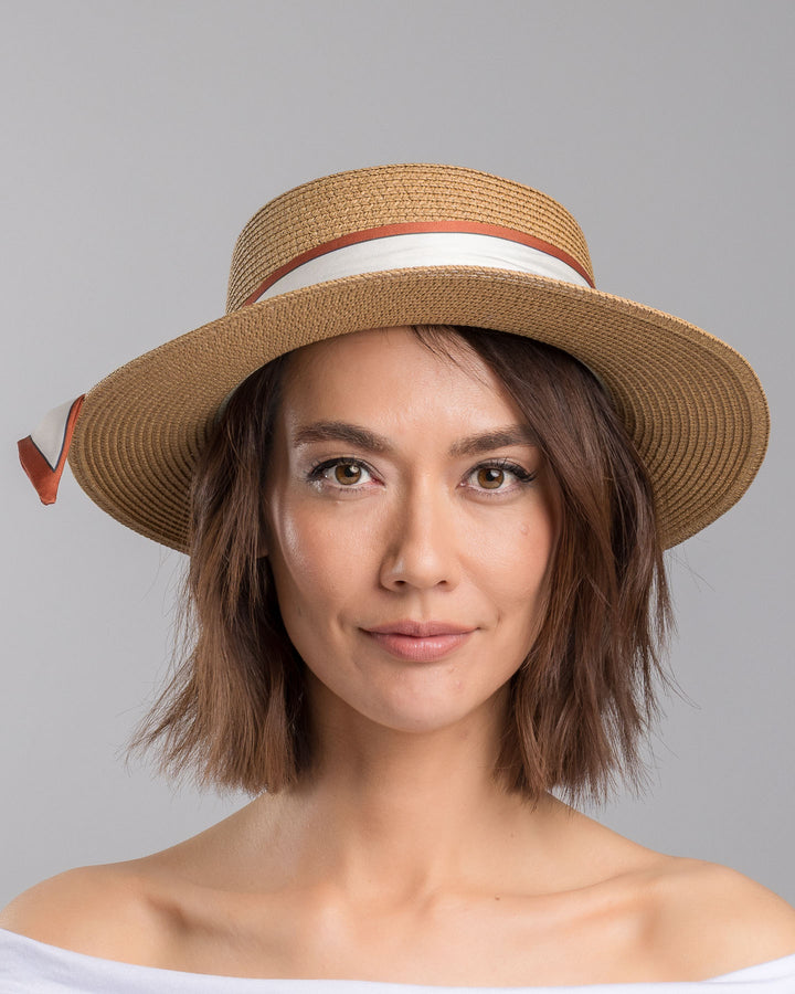 The model is showcasing the front side close up of the natural woven straw boater hat with a wide brim, wrapped in a cream and rust silk ribbon and finished with a tied scarf featuring an illustrated female portrait detail. Design and made by Merve Bayindir millinery.