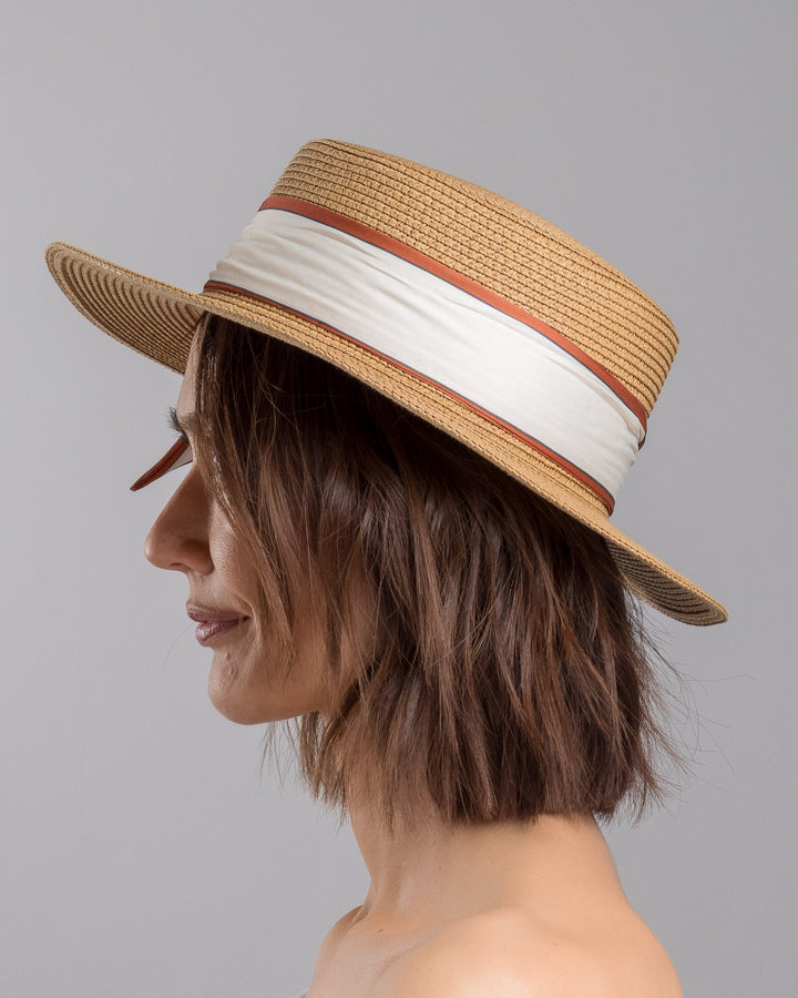 The model is showcasing the left side close up of the natural woven straw boater hat with a wide brim, wrapped in a cream and rust silk ribbon and finished with a tied scarf featuring an illustrated female portrait detail. Design and made by Merve Bayindir millinery.