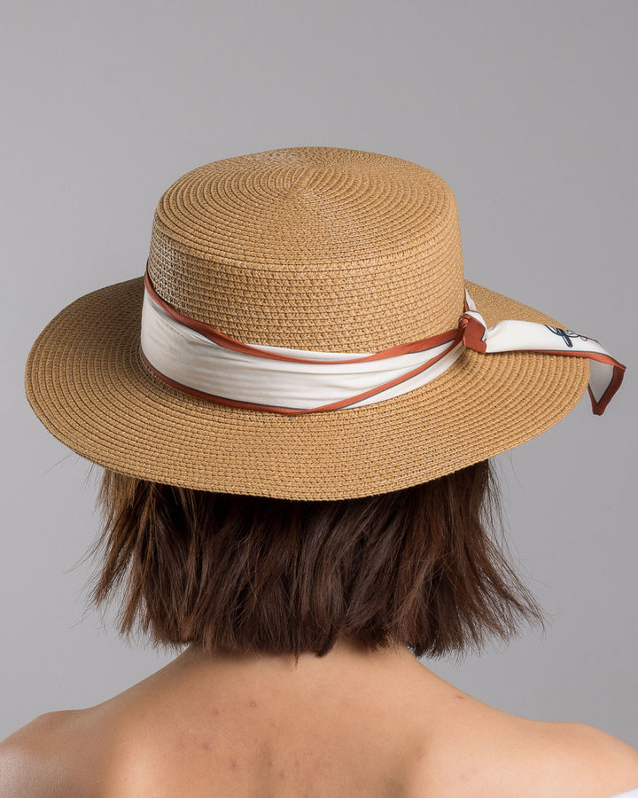 The model is showcasing the back side close up of the natural woven straw boater hat with a wide brim, wrapped in a cream and rust silk ribbon and finished with a tied scarf featuring an illustrated female portrait detail. Design and made by Merve Bayindir millinery.